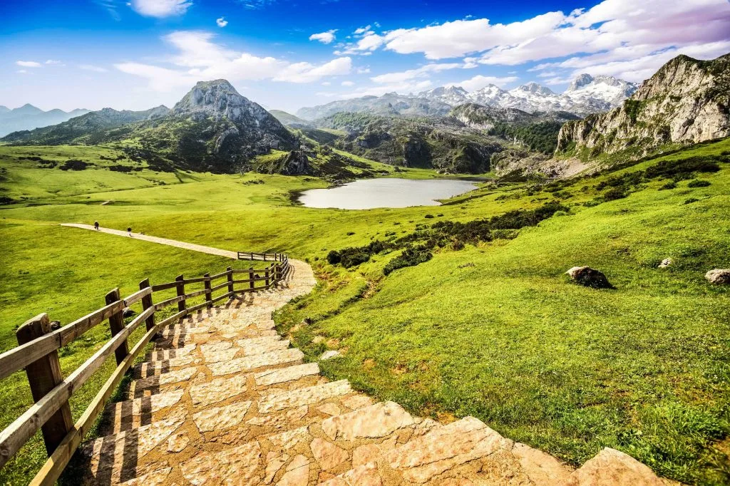 Paisaje montañoso en los Picos de Europa, con un sendero de piedra que desciende hacia un lago rodeado de vegetación verde y montañas. El cielo está despejado con algunas nubes. Ideal para el senderismo y la naturaleza.