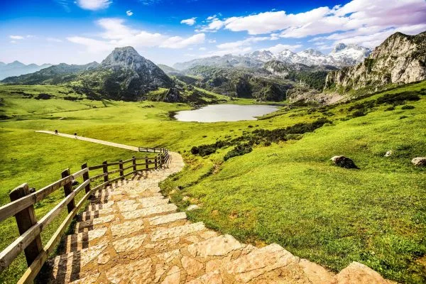 Paisaje montañoso en los Picos de Europa, con un sendero de piedra que desciende hacia un lago rodeado de vegetación verde y montañas. El cielo está despejado con algunas nubes. Ideal para el senderismo y la naturaleza.