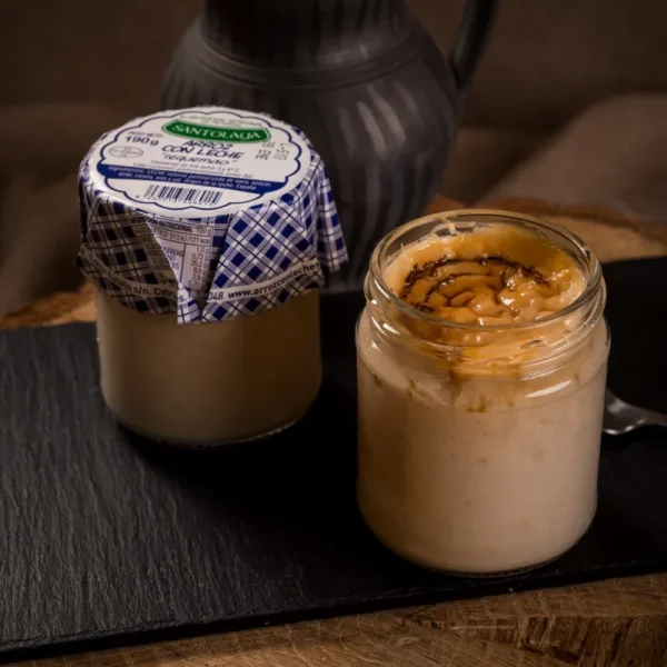 Dos frascos de postre en un plato de piedra. A la izquierda, un frasco con tapita de cuadros azules que contiene arroz con leche. A la derecha, otro frasco con una deliciosa capa de caramelo en la parte superior. Fondo neutral y texturizado.