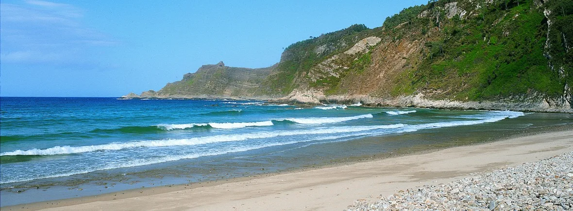Playa tranquila con olas suaves y acantilados verdes al fondo, ideal para disfrutar de la naturaleza y actividades acuáticas.