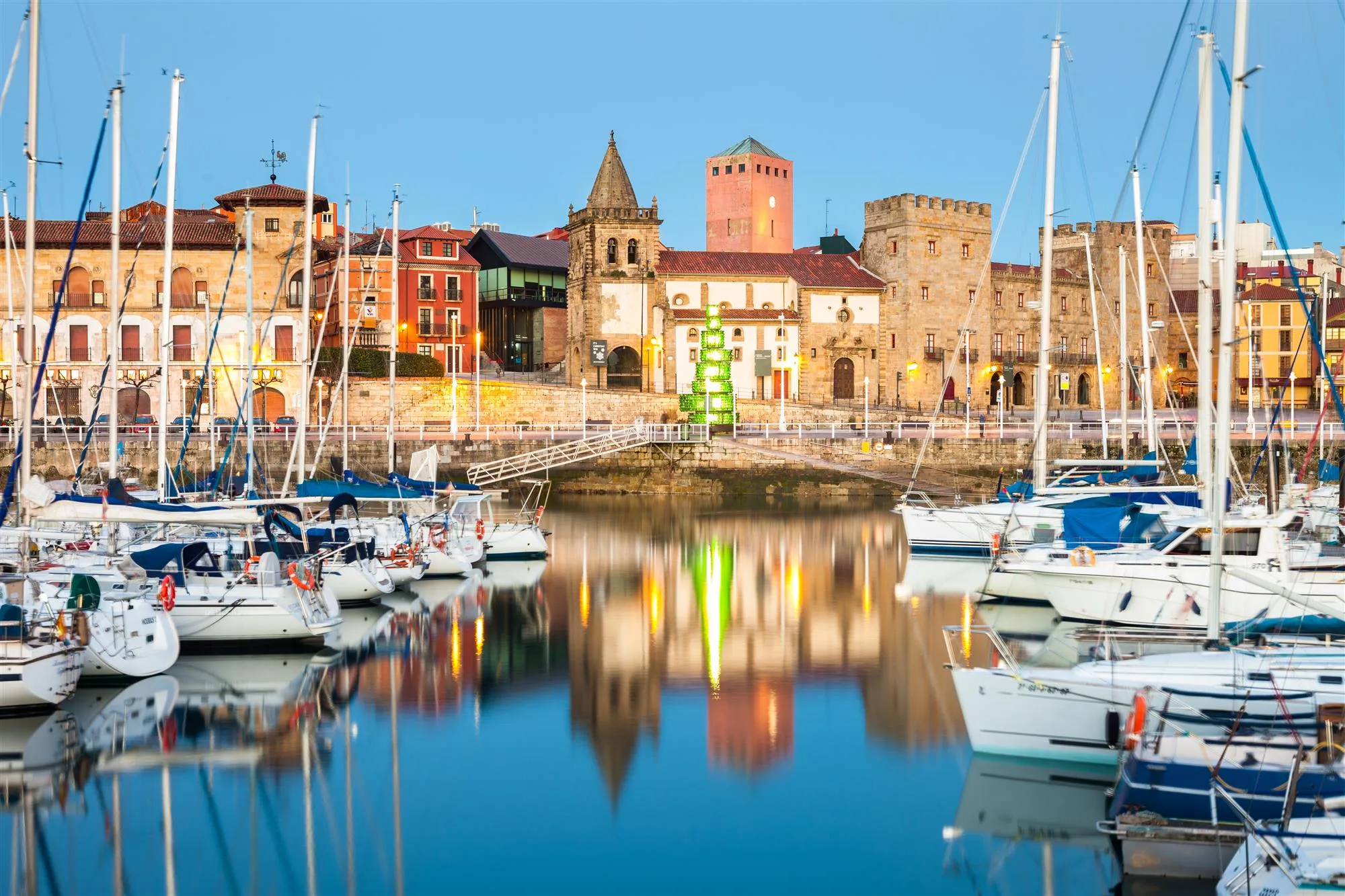 Puerto de un pueblo costero con barcos de vela amarrados, edificios históricos y reflejos en el agua al atardecer.