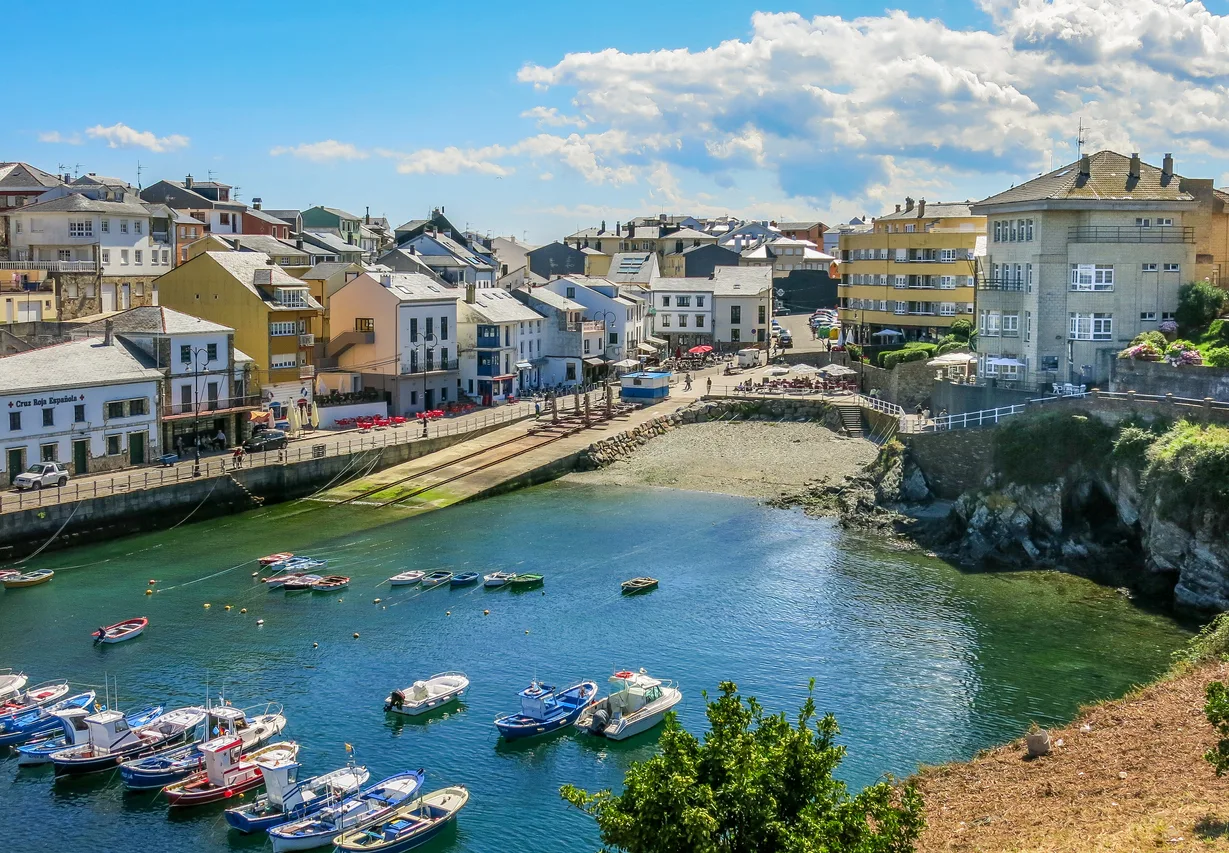 Vista panorámica de un puerto en una localidad costera de España, con barcos pesqueros en el agua y edificios coloridos a lo largo del muelle. El cielo es azul con algunas nubes, creando un ambiente soleado y atractivo.