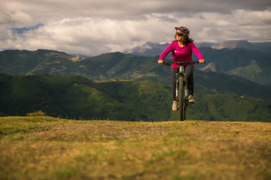 Mujer montando una bicicleta de montaña en un paisaje montañoso, con nubes y vegetación verde de fondo, disfrutando de la aventura al aire libre.