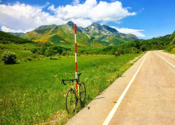 Bicicleta de carretera apoyada en un poste de señalización, con un paisaje montañoso y verde al fondo, bajo un cielo azul con nubes. Ideal para ciclismo y actividades al aire libre.