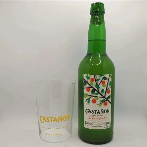 Botella de sidra natural Castañón de color verde con etiqueta decorativa y vaso transparente con el logo de Castañón. Ideal para disfrutar de bebidas tradicionales.