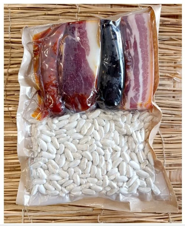 Paquete de carne curada y frijoles blancos, con cuatro tipos de tocino en la parte superior y frijoles en la parte inferior, todo envasado al vacío. Ideal para preparar platos tradicionales.