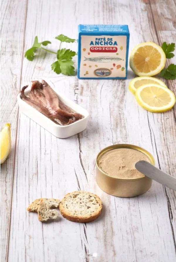 Paté de anchoa en lata junto a filetes de anchoa en un plato blanco, rodajas de limón y pan tostado sobre una mesa de madera. Ideal para aperitivos y recetas gourmet.