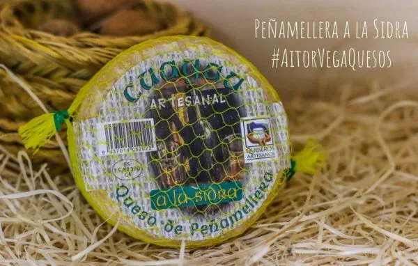 Queso de Peñamellera a la Sidra, envuelto en malla amarilla, sobre un fondo de paja. Producto artesanal de Queseros Artesanos, ideal para acompañar con sidra.
