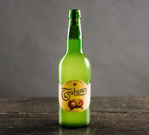 Botella de sidra natural Crabanco, con etiqueta que muestra dos manzanas, sobre fondo oscuro y superficie de madera. Ideal para resaltar la tradición de la sidra asturiana.